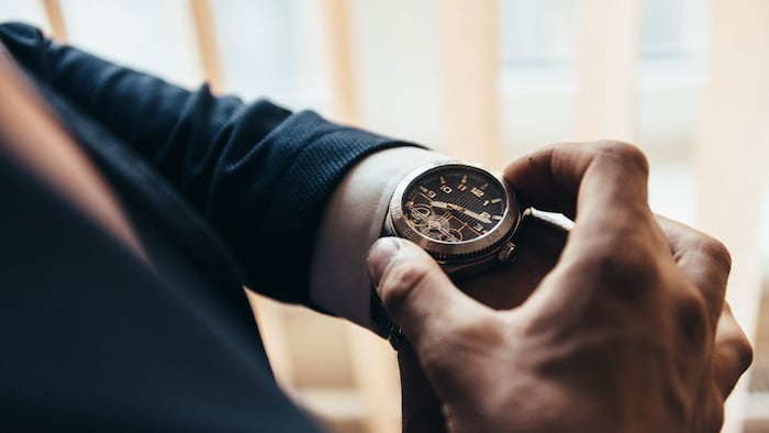 a stylish mechanical watch on the arm of a man dressed in a blue jacket with a white shirt that watches the time on the clock holding the clock by hand